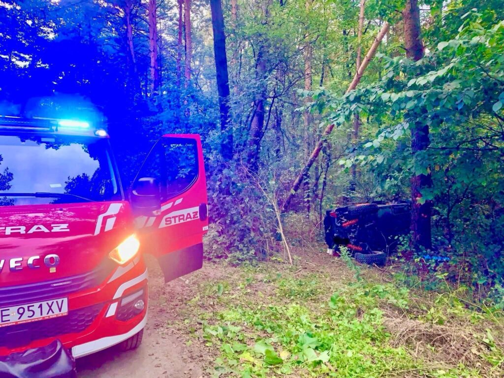 Czerwony wóz strażacki z włączonymi sygnałami świetlnymi stoi na leśnej drodze obok przewróconego pojazdu.