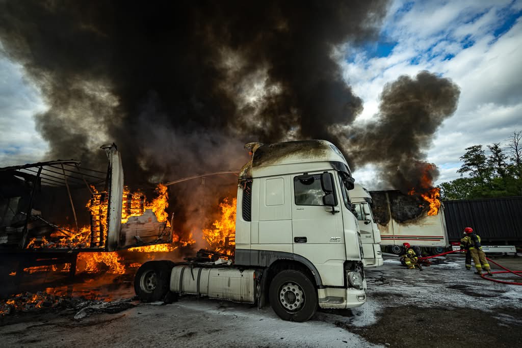 Białe ciężarówki płoną w dymie, a strażacy gaszą ogień przy użyciu węży na terenie przemysłowym.