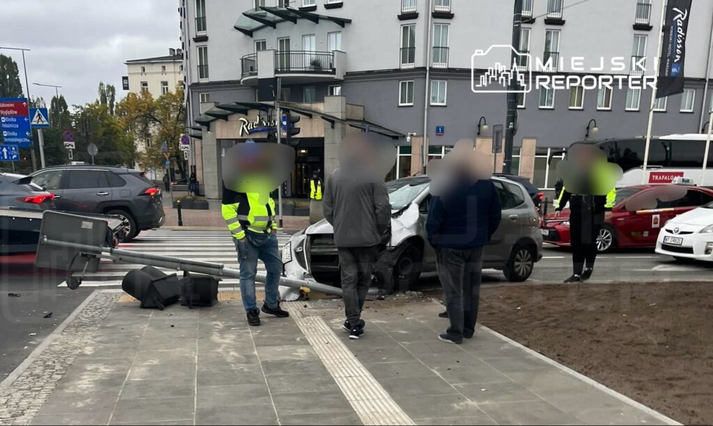 Wypadek drogowy na skrzyżowaniu, z uszkodzonym samochodem i przewróconym słupem. Policja i świadkowie zbierają informacje.