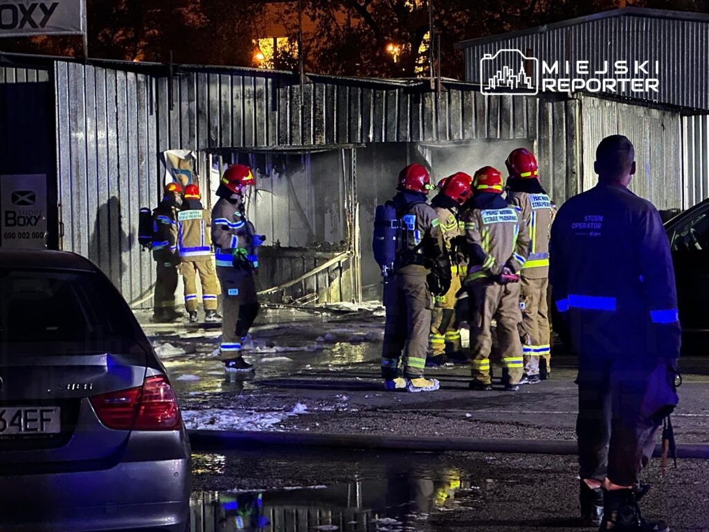 Grupa strażaków w hełmach omawia działania ratunkowe przy palącym się budynku, obok widać samochód.