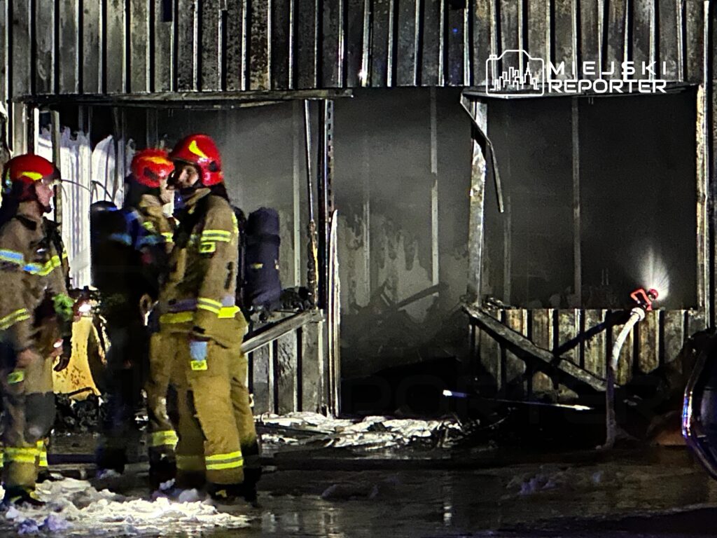 Grupa strażaków w hełmach rozmawia obok zniszczonego budynku po pożarze, w tle widać wodę i sprzęt gaśniczy.