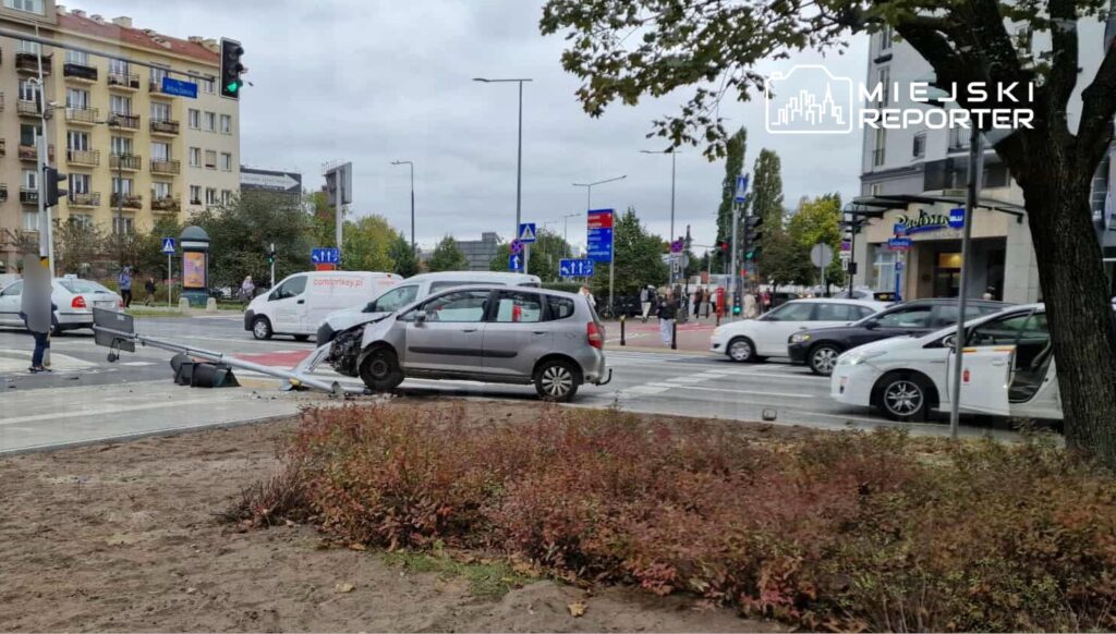 Uszkodzony samochód osobowy stoi na skrzyżowaniu, obok przewrócony znak drogowy, a w tle widać inne pojazdy.