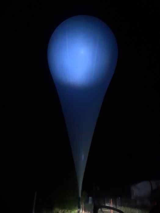 Niebieski balon meteorologiczny unosi się w nocy, oświetlając otoczenie delikatnym blaskiem.