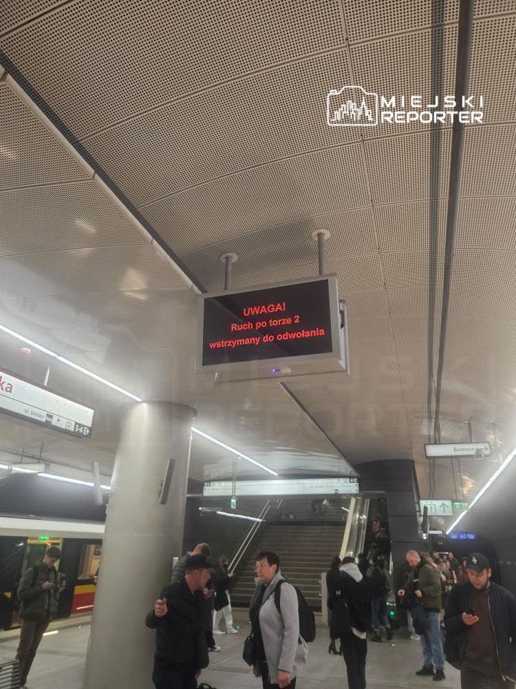 Pasażerowie na stacji metra czekają obok wyświetlacza z komunikatem: "UWAGA! Ruch po torze 2 wstrzymany do odwołania".