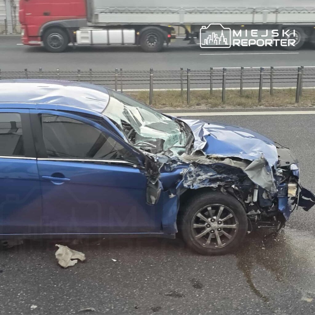Niebieski samochód osobowy z uszkodzoną przednią częścią stoi na poboczu drogi, obok przejeżdżających ciężarówek.