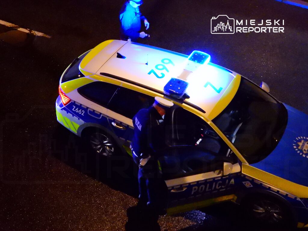 Policjant w mundurze rozmawia z innym funkcjonariuszem obok radiowozu z włączonymi sygnałami świetlnymi.