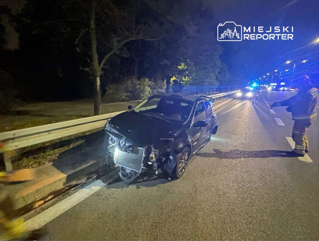 Czarny samochód osobowy uszkodzony po kolizji, zatrzymany przy barierce na drodze, obok strażak w mundurze.