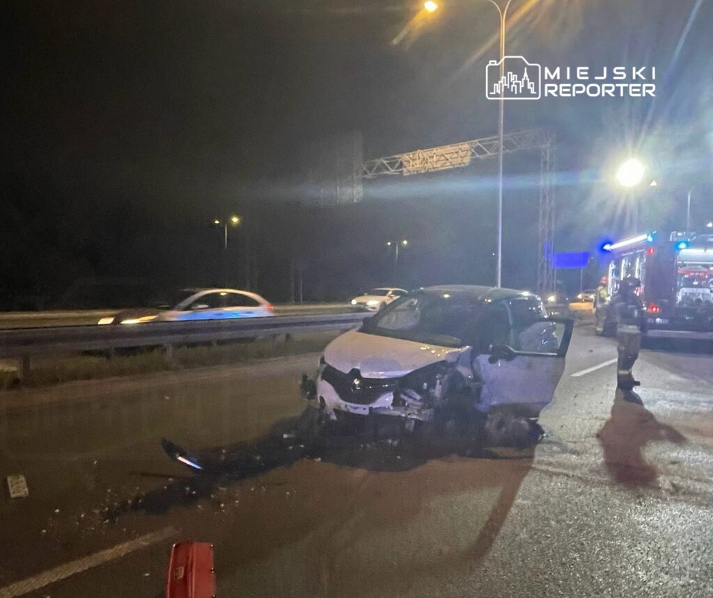 Uszkodzony samochód osobowy na drodze, obok strażaków oraz wozu strażackiego w nocy.