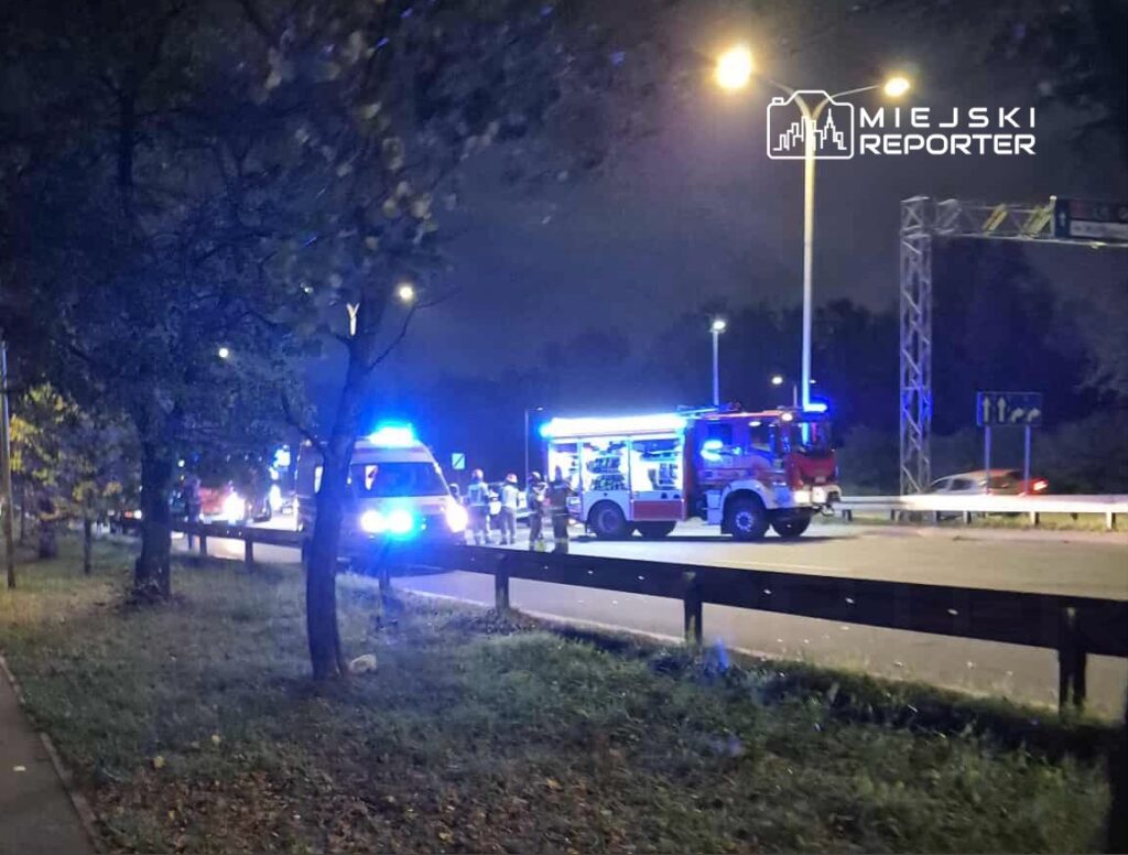 Na drodze nocą, z niebieskimi światłami, strażacy pracują przy samochodzie ratunkowym obok bariery ochronnej.