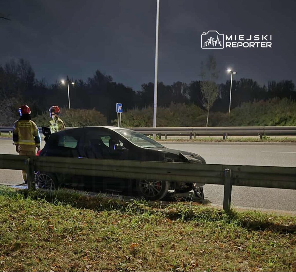 Dwóch strażaków w kaskach rozmawia obok uszkodzonego samochodu na poboczu drogi nocą.