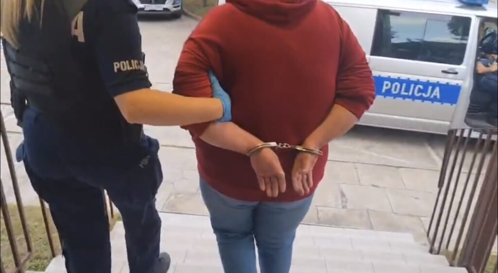 Kobieta w czerwonym swetrze jest prowadzona przez policjantkę w mundurze, ręce ma skute w kajdankach.