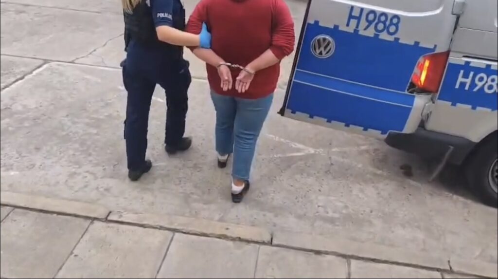 Kobieta w czerwonym swetrze jest zatrzymywana przez policjantkę w niebieskim mundurze na ulicy.
