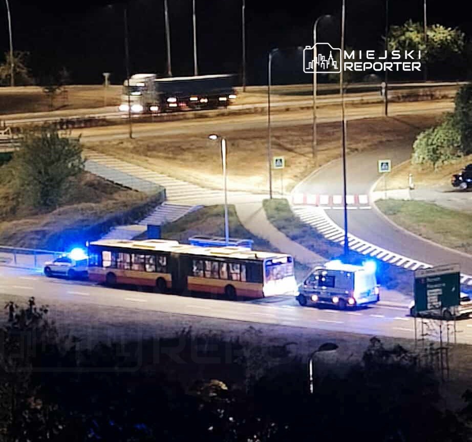 Autobus z włączonymi światłami awaryjnymi zatrzymany na poboczu drogi, obok niego radiowóz policyjny z niebieskimi sygnalizatorami.