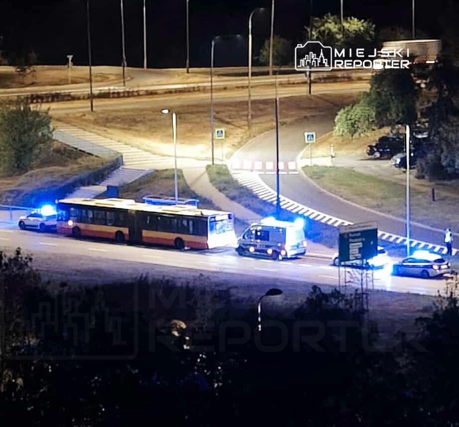 Autobus z włączonymi światłami awaryjnymi zatrzymany na drodze, otoczony przez radiowozy policyjne z niebieskimi sygnałami świetlnymi.