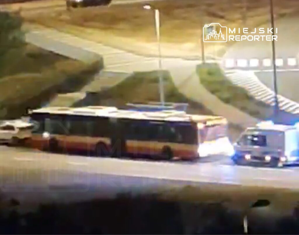 Autobus komunikacji miejskiej zatrzymany na skrzyżowaniu, obok niego radiowóz z włączonymi sygnałami świetlnymi.