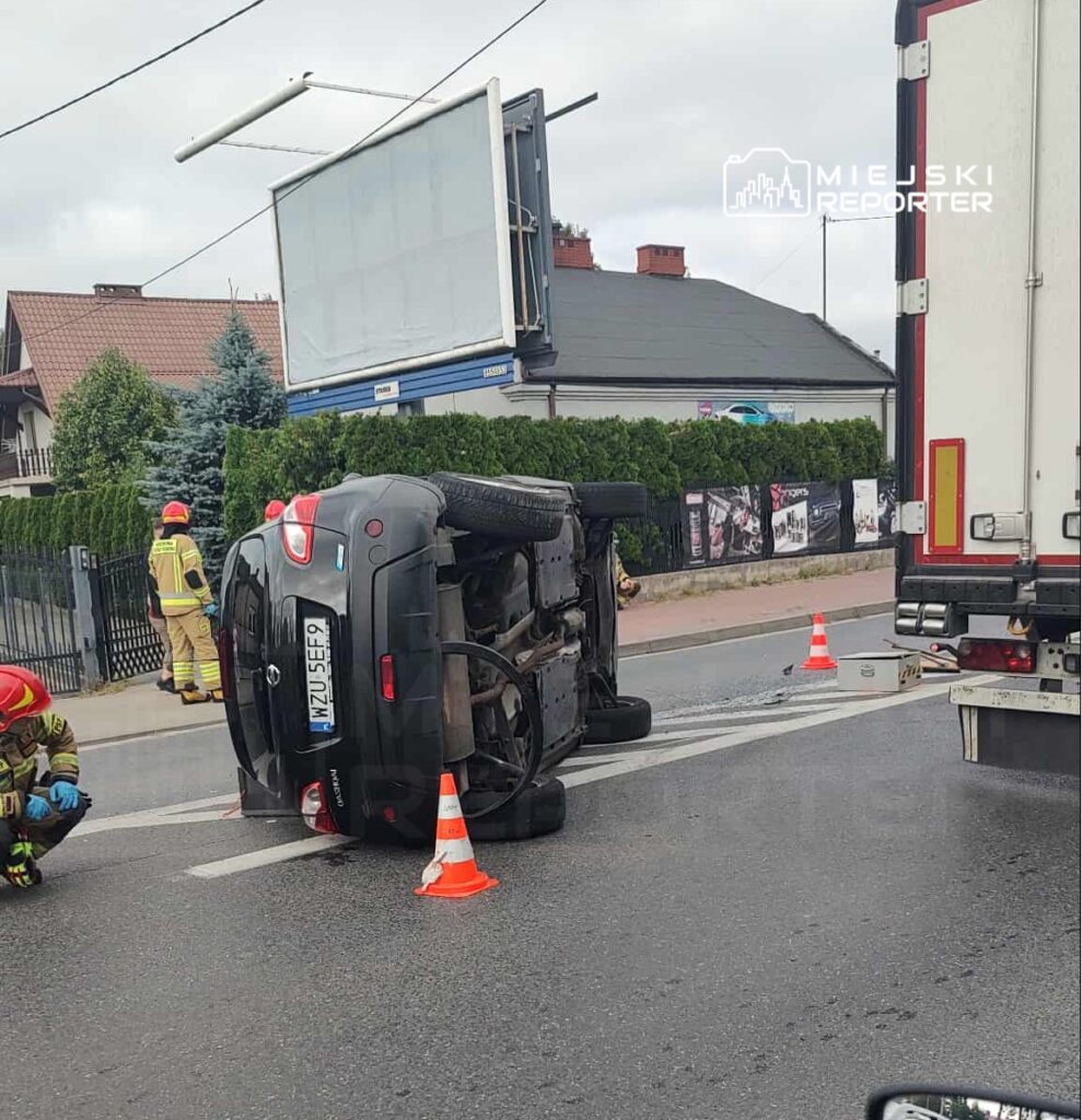 Samochód osobowy leży na boku na jezdni, a w pobliżu pracują strażacy, zabezpieczając miejsce zdarzenia.