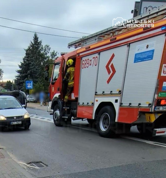 Strażak w hełmie wchodzi do wozu strażackiego na ulicy, obok zatrzymany samochód osobowy.
