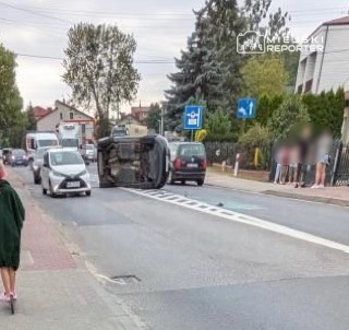 Na drodze leży przewrócony samochód, obok niego stoją inne pojazdy, a w tle widoczni przechodnie.