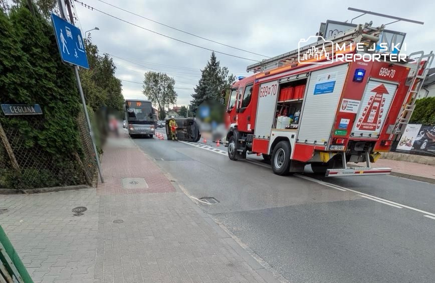 Czerwony wóz strażacki zatrzymany przy drodze, obok przewrócony autobus oraz pracujący strażacy w kamizelkach odblaskowych.