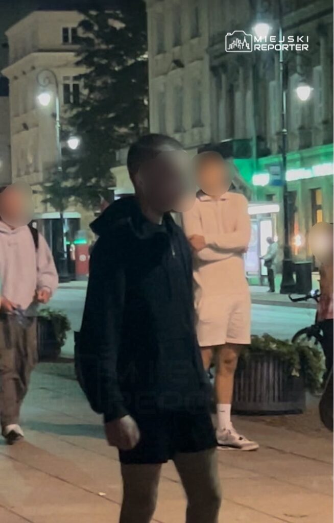 Grupa młodych ludzi spacerujących po oświetlonej ulicy, w tle widać budynki i lampy uliczne.