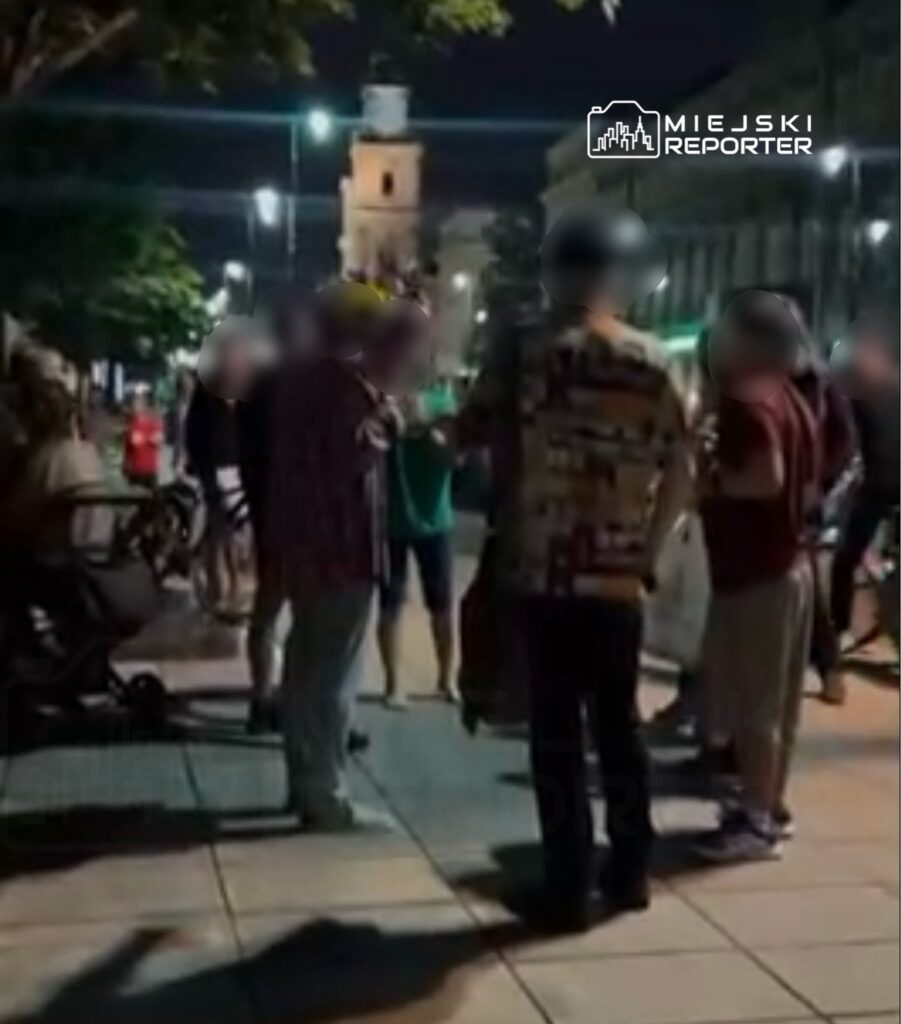 Grupa ludzi rozmawia na chodniku nocą, w tle widoczny budynek z wieżą zegarową.