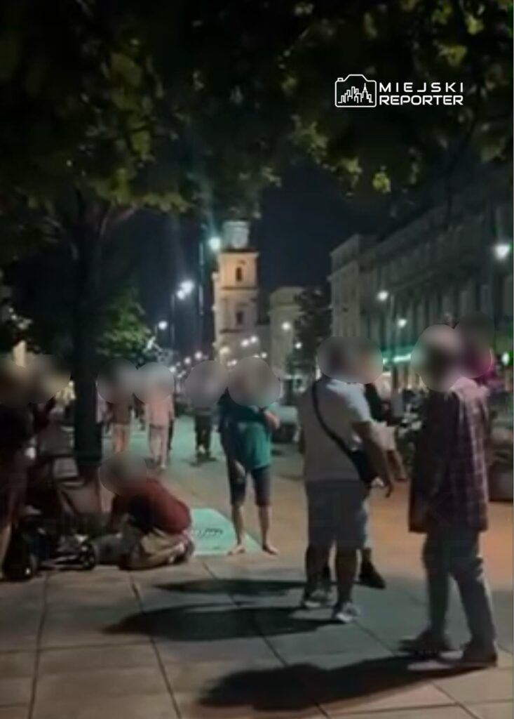 Grupa ludzi rozmawia na oświetlonej ulicy nocą, w tle widoczny zegar na wieży.