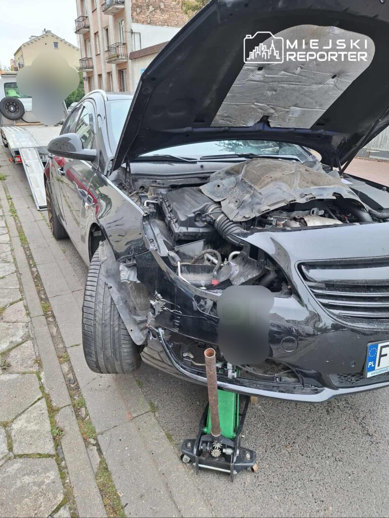 Czarny samochód osobowy z otwartą maską i uszkodzeniami, unieruchomiony na chodniku, podparty podnośnikiem.