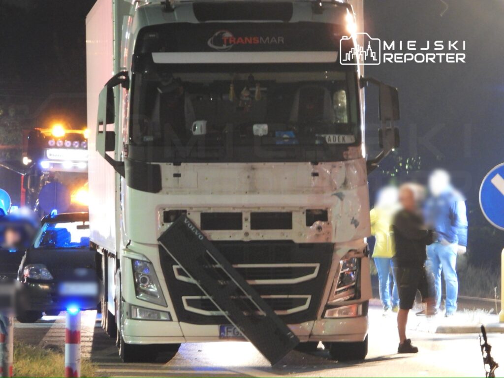 Ciężarówka z naczepą zatrzymana na drodze, obok stoją policjanci oraz kierowca osobowego auta. W tle widoczne światła radiowozu.
