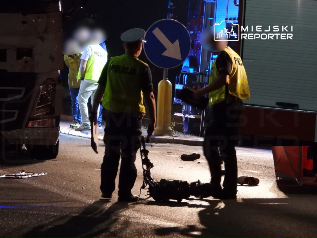 Policjanci prowadzą dochodzenie na miejscu wypadku drogowego, obok uszkodzonego pojazdu i znaku drogowym.