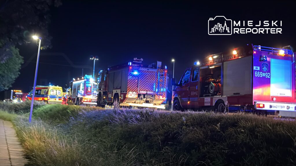 Zespół ratowniczy z wozami strażackimi i ambulansami pracuje na miejscu wypadku nocą, przy oświetlonej drodze.