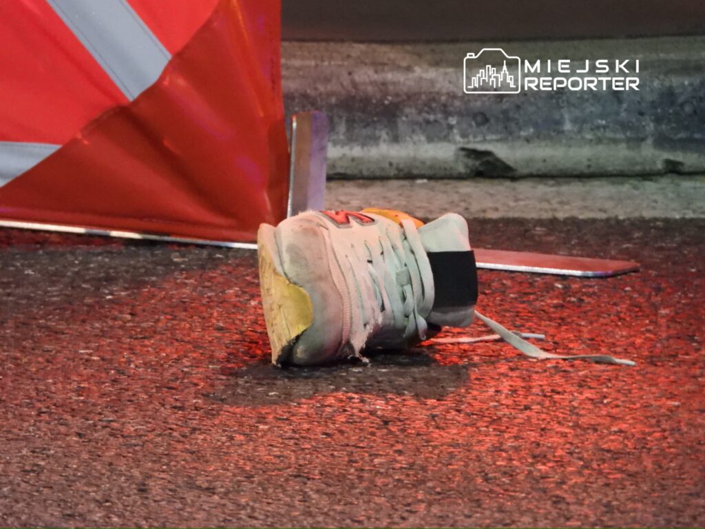 Zniszczony but sportowy leży na asfalcie obok bariery drogowej, w tle odbicia świateł.