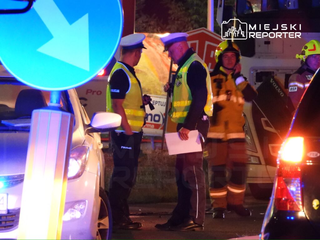 Dwóch policjantów w odblaskowych kamizelkach rozmawia przy drodze, obok stoją strażacy w mundurach.