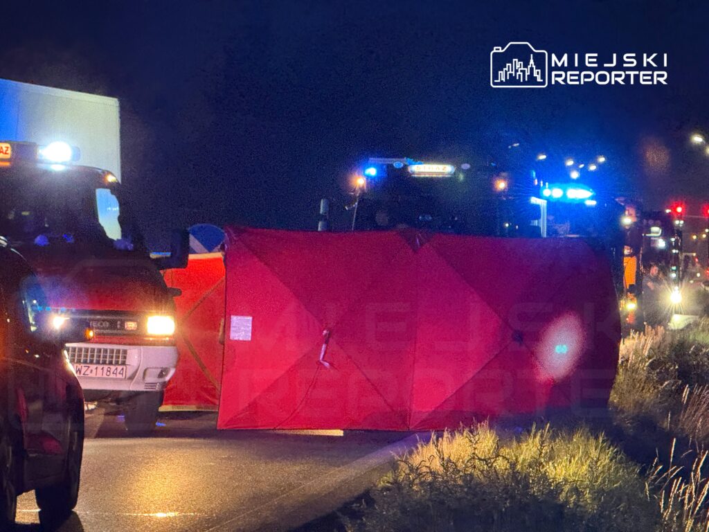 Na drodze stoi czerwony namiot, a w tle widoczne są pojazdy służb ratunkowych z włączonymi sygnałami świetlnymi.