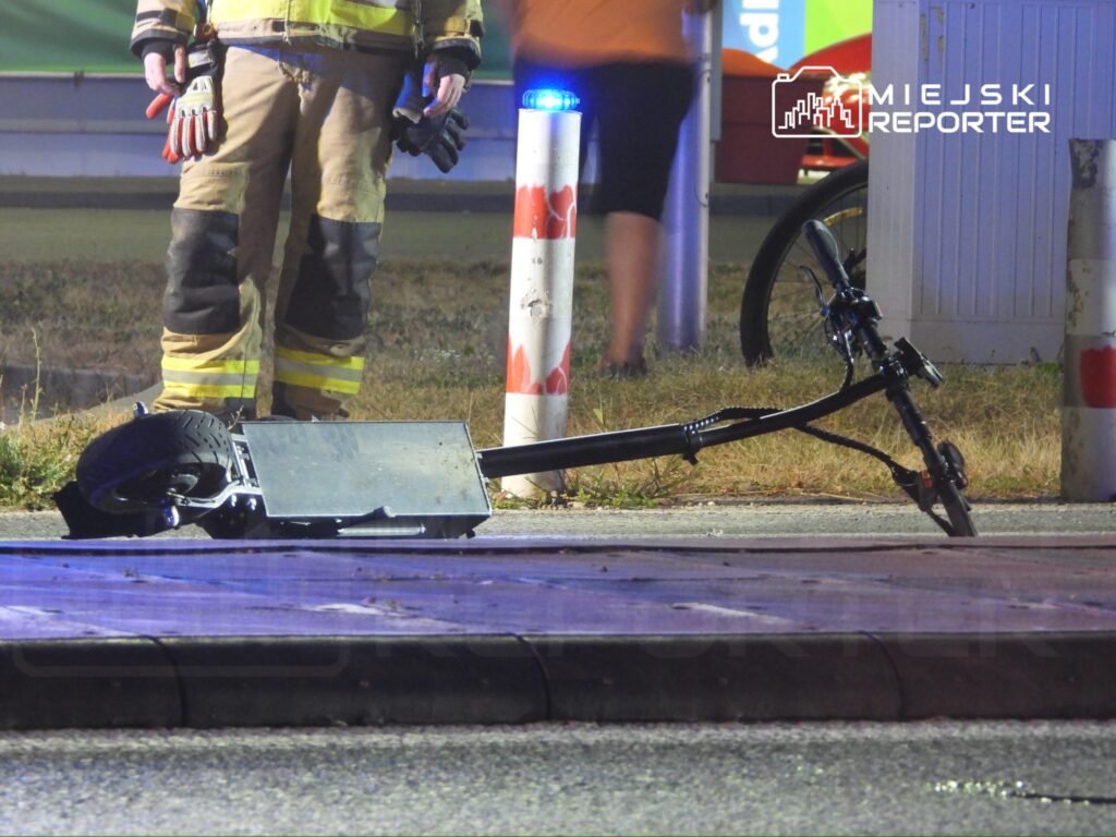 Strażak w mundurze stoi obok uszkodzonej hulajnogi elektrycznej leżącej na jezdni.