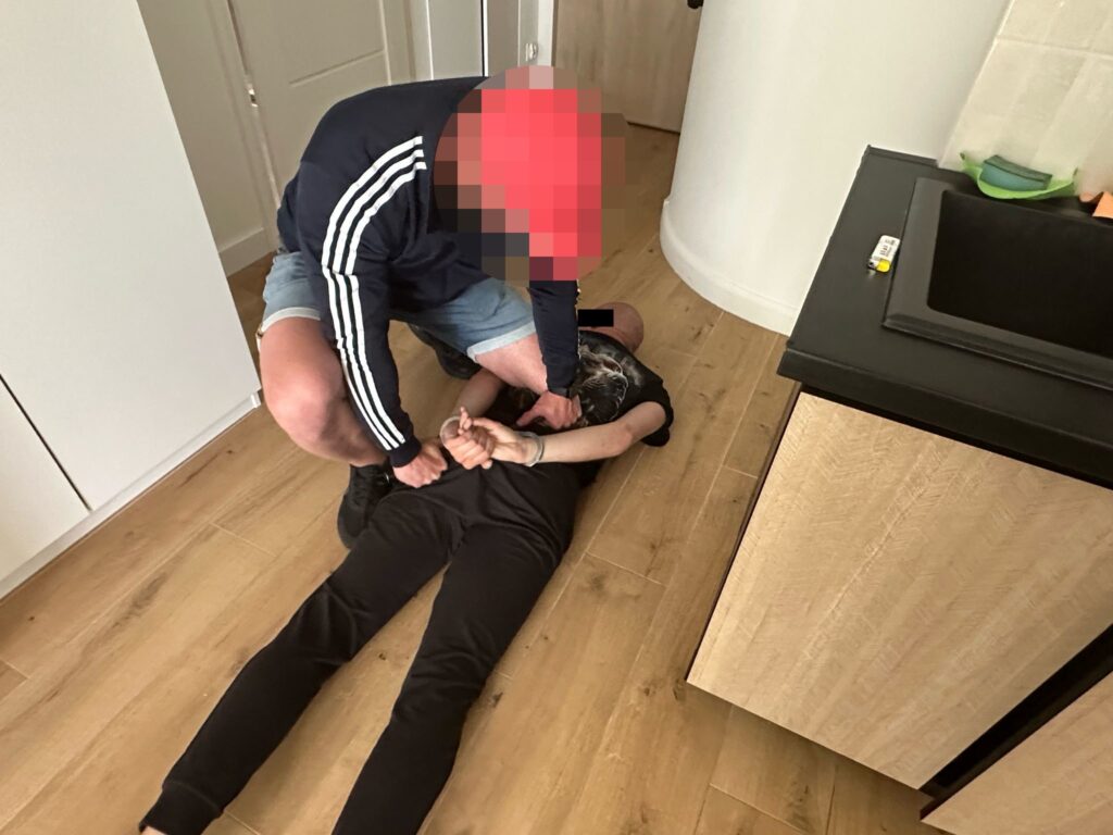 Mężczyzna w sportowej odzieży przytrzymuje leżącego mężczyznę na podłodze w nowoczesnym wnętrzu mieszkania.