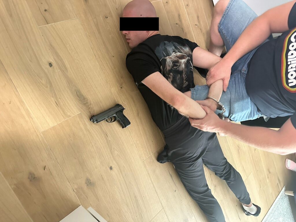 Mężczyzna leżący na podłodze z rękami związanymi kajdankami, obok niego leży pistolet.