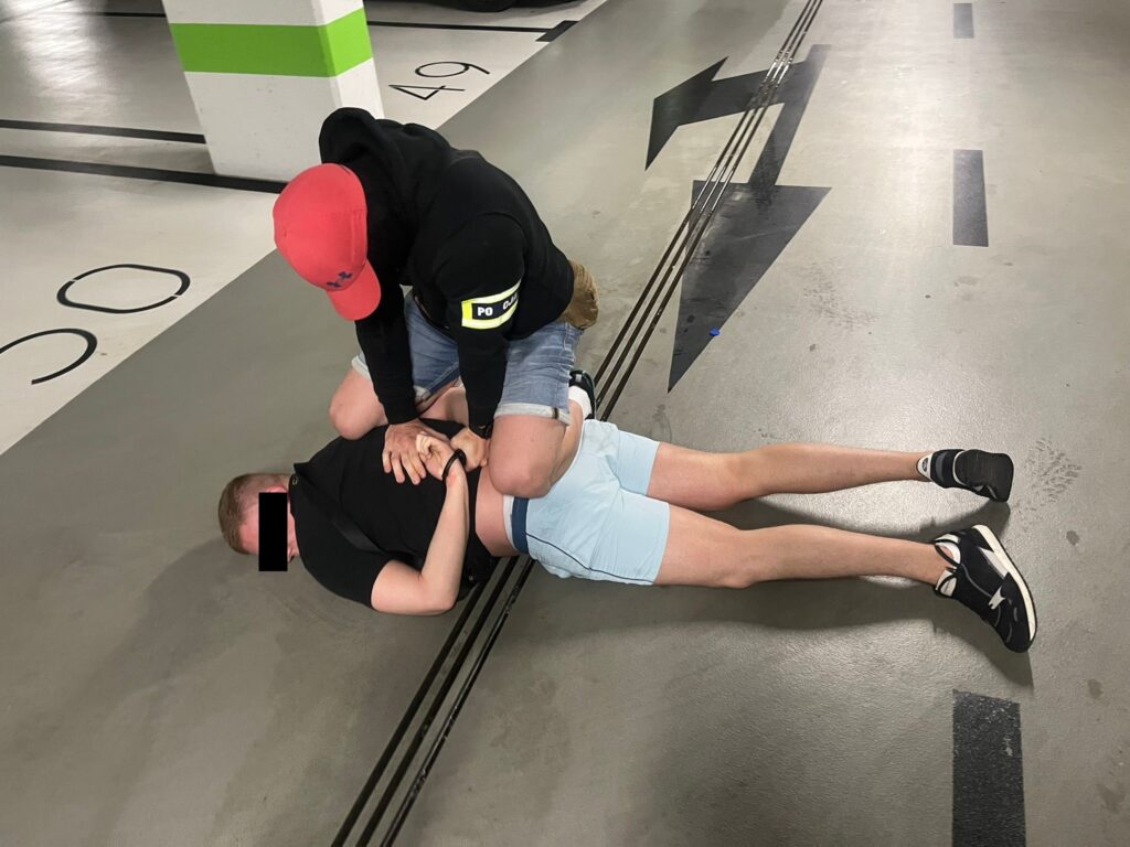 Mężczyzna w czarnej bluzie i czerwonej czapce przytrzymuje innego mężczyznę leżącego na podłodze w parkingu.