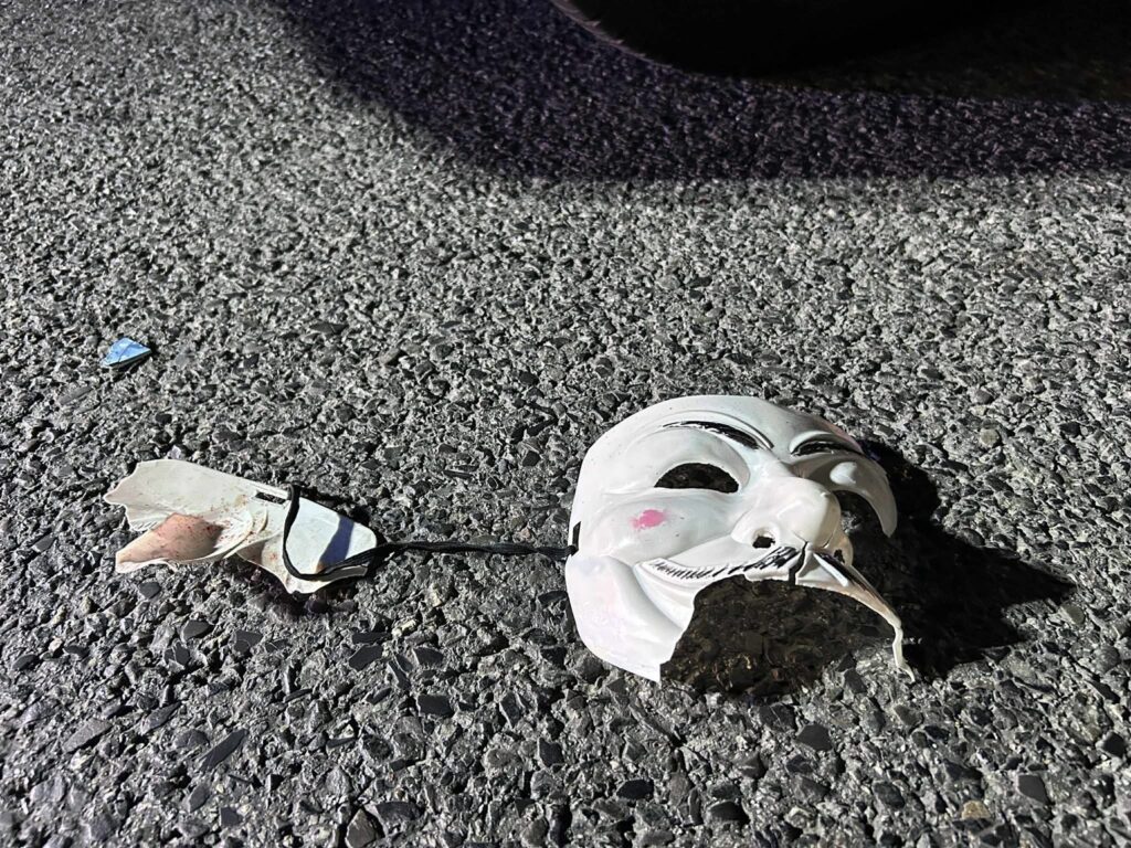 Złamana maska leży na asfaltowej nawierzchni obok fragmentu lustra.