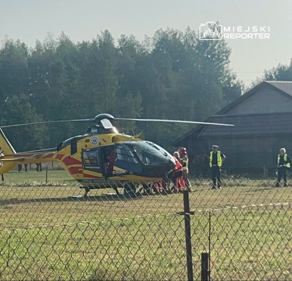 Lądowanie śmigłowca ratunkowego na otwartym terenie, obok zespołu ratowników i osób w kamizelkach odblaskowych.