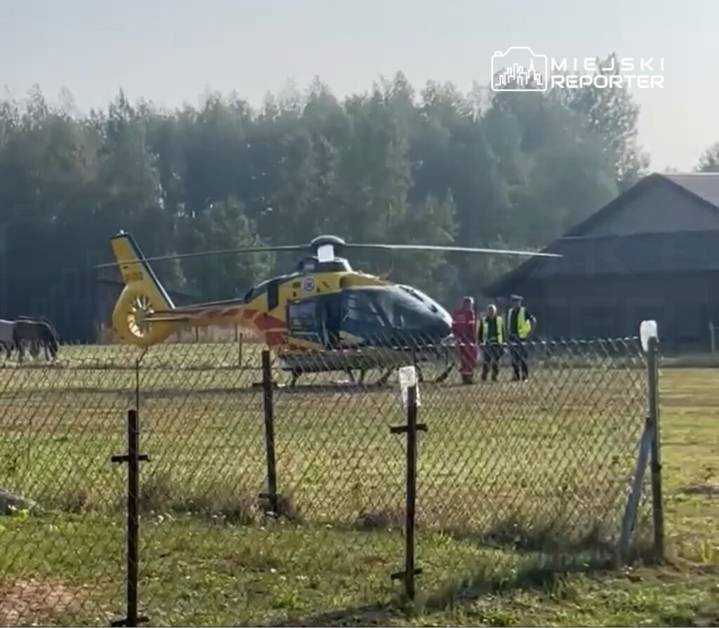 Helikopter lądował na łące, obok stoją konie, a w pobliżu grupka ludzi w odzieży ochronnej.