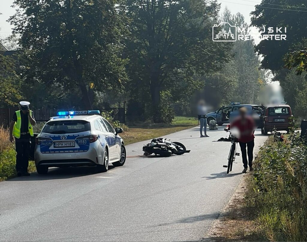 Policjant w kamizelce odblaskowej rozmawia przez telefon obok przewróconego motocykla na drodze, a rowerzysta prowadzi rower.