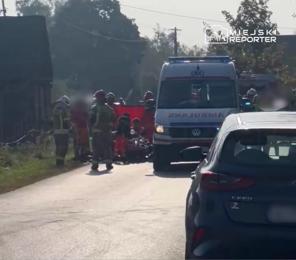 Grupa ratowników i strażaków udziela pomocy poszkodowanemu na drodze, obok stoi karetka.