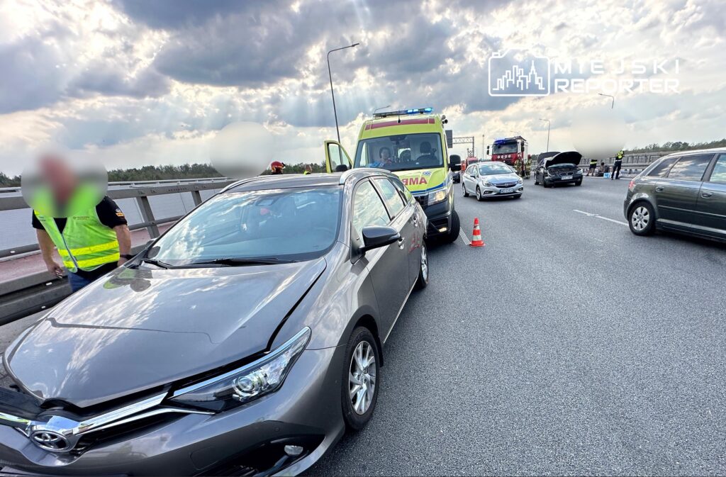 Wypadek drogowy na moście, z uszkodzonym samochodem osobowym i pojazdem ratunkowym w tle.