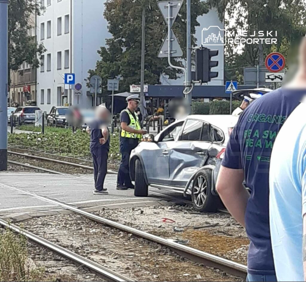 Policjant rozmawia z kierowcą uszkodzonego samochodu na skrzyżowaniu obok torów tramwajowych.