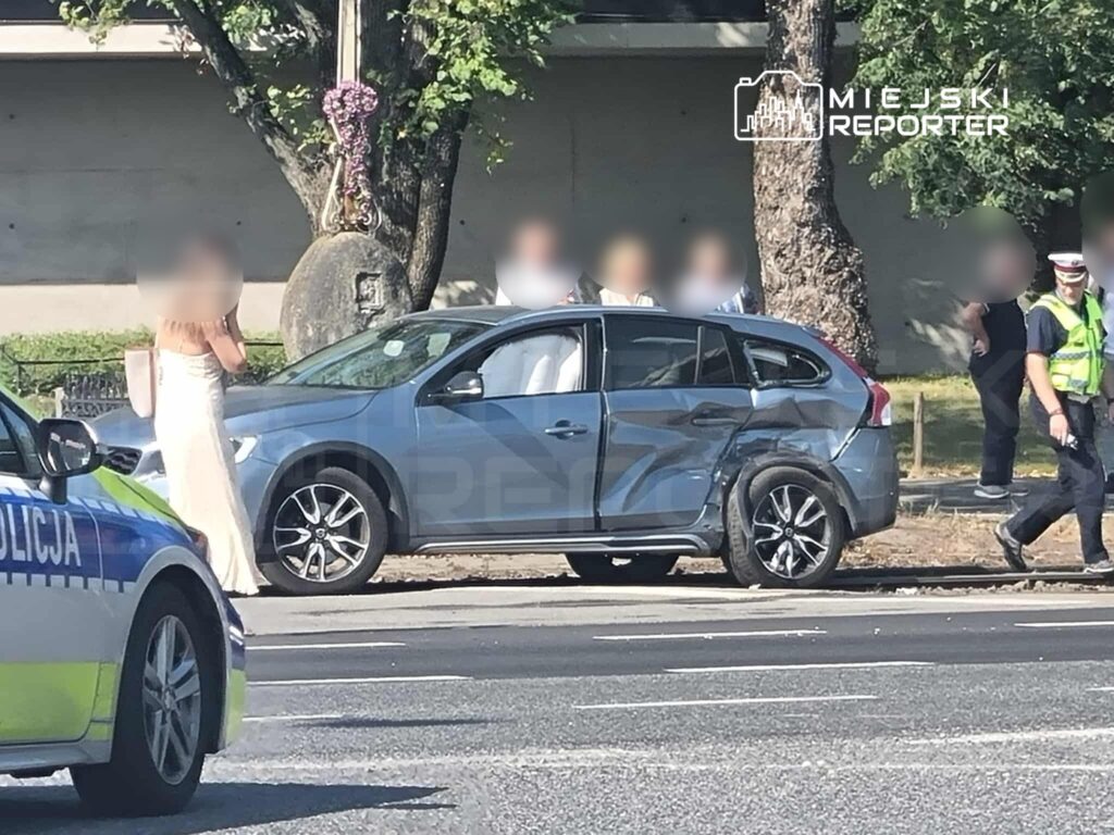 Kobieta w sukni wieczorowej rozmawia przez telefon obok uszkodzonego samochodu, w tle interweniujący policjanci.