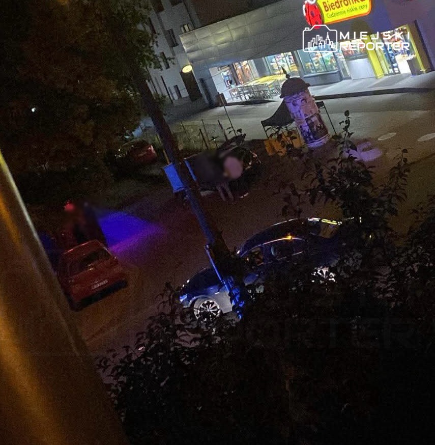 Grupa osób stoi obok samochodu na ulicy, w tle widoczny sklep spożywczy oświetlony neonami.