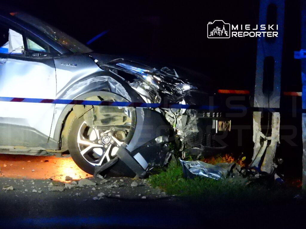 Uszkodzony samochód osobowy z wgniecioną stroną, zderzony z słupem, na drodze nocą, otoczony taśmą policyjną.
