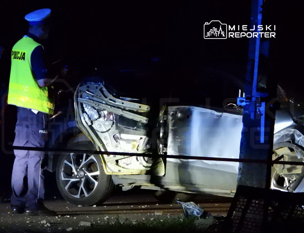Policjant w kamizelce odblaskowej bada uszkodzony samochód w nocy, przy drodze, otoczony przez taśmy zabezpieczające.