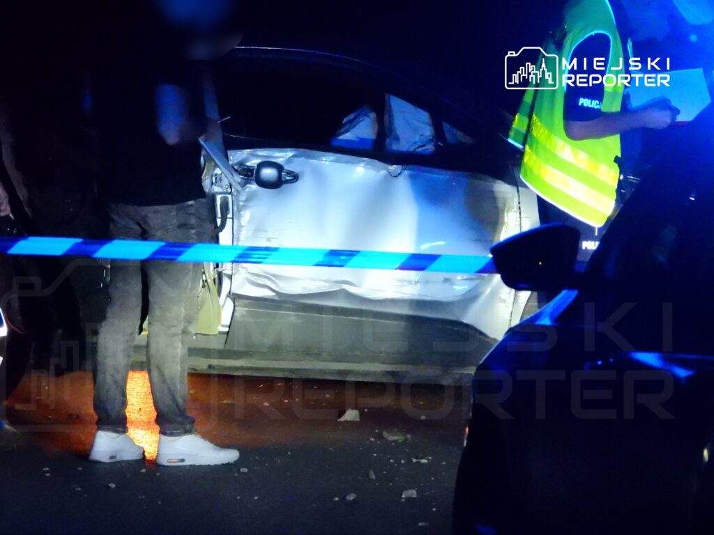Mężczyzna w czarnej koszulce stoi obok uszkodzonego samochodu, podczas gdy policjant w kamizelce odblaskowej notuje informacje.