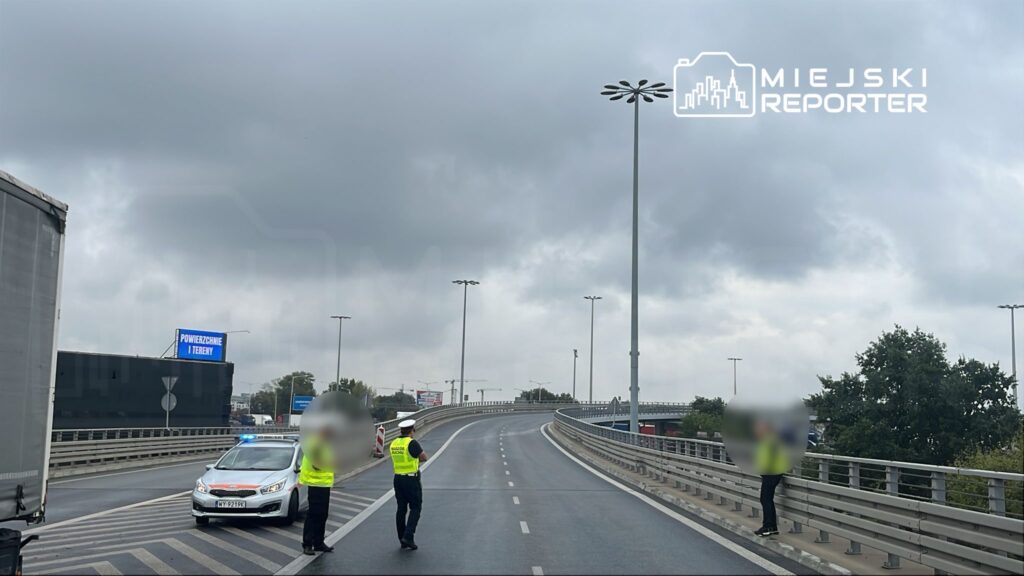 Policjanci w kamizelkach odblaskowych kierują ruchem na drodze, obok zaparkowany radiowóz z włączonymi światłami.